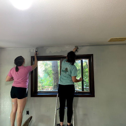 two students, standing on a chair and short ladder, paint a wall near a ceiling of a home