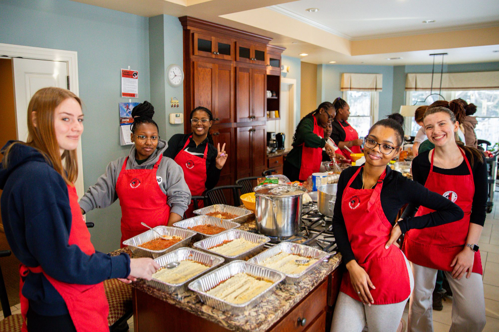students in red aprons gathered around kitchen island with food in catering dishes are pictured