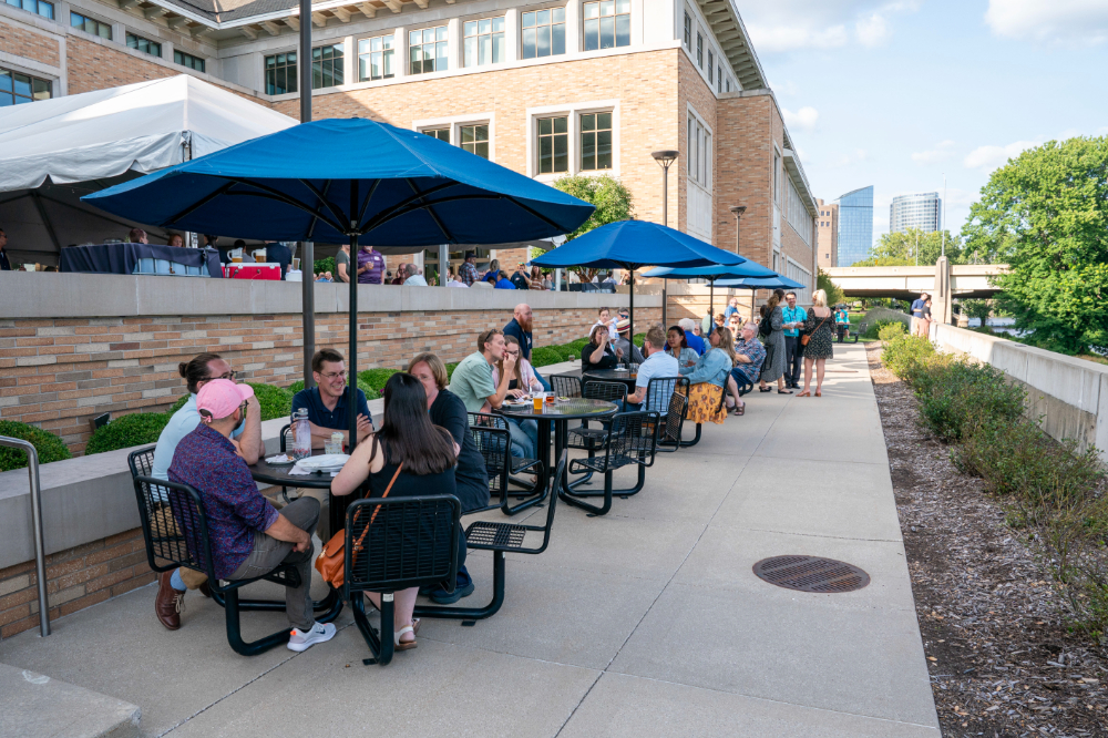 blue umbrellas line the sidewalk between the Seidman Center and the Grand River, people sitting at black tables under the umbrellas