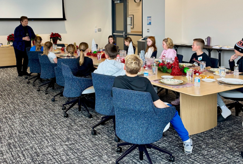 Cindy Brown stands in front of a long conference table, young students seated at table with plates and silverware and glasses set on the table