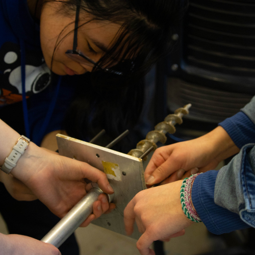 three people holding a mechanical device; two sets of hands on this metal while one student is seen on the left