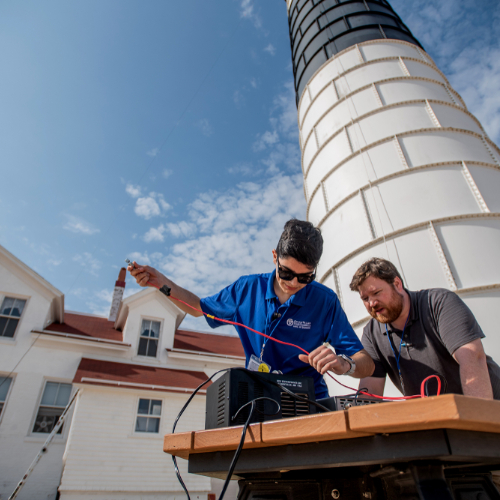 in front of a lighthouse, two people work at a table fixing wires into a ham radio