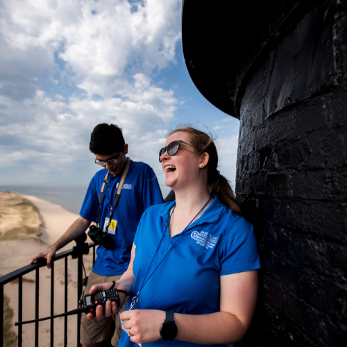two people holding a wire on a balcony of a lighthouse with sand dunes and Lake Michigan in background