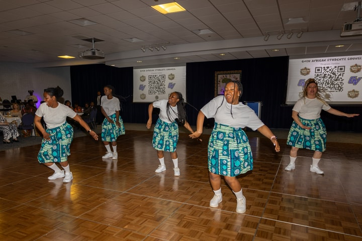 five dancers in white t-shirts and colorful green shorter skirts dance on a wooden stage
