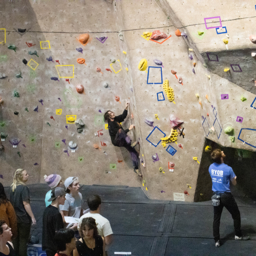 Two people on climbing wall, one person watching from below in blue shirt; crowd at left