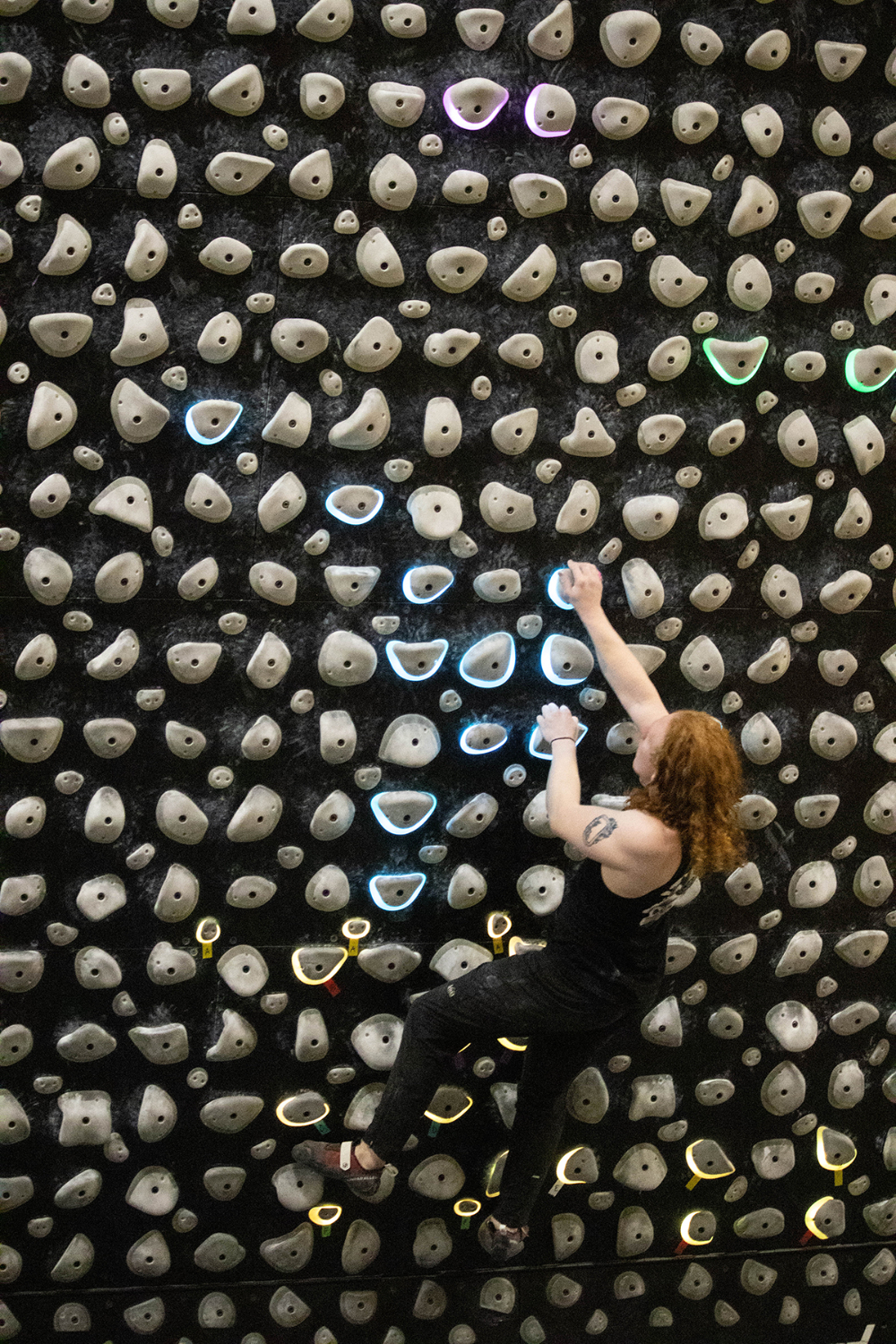 student with long red hair in black tshirt and pants climbs the climbing wall, some rocks are lit behind