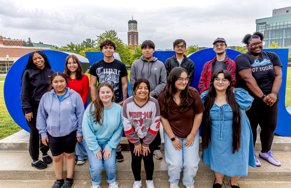 two rows of students standing in front of the GVSU letters, carillon tower in background