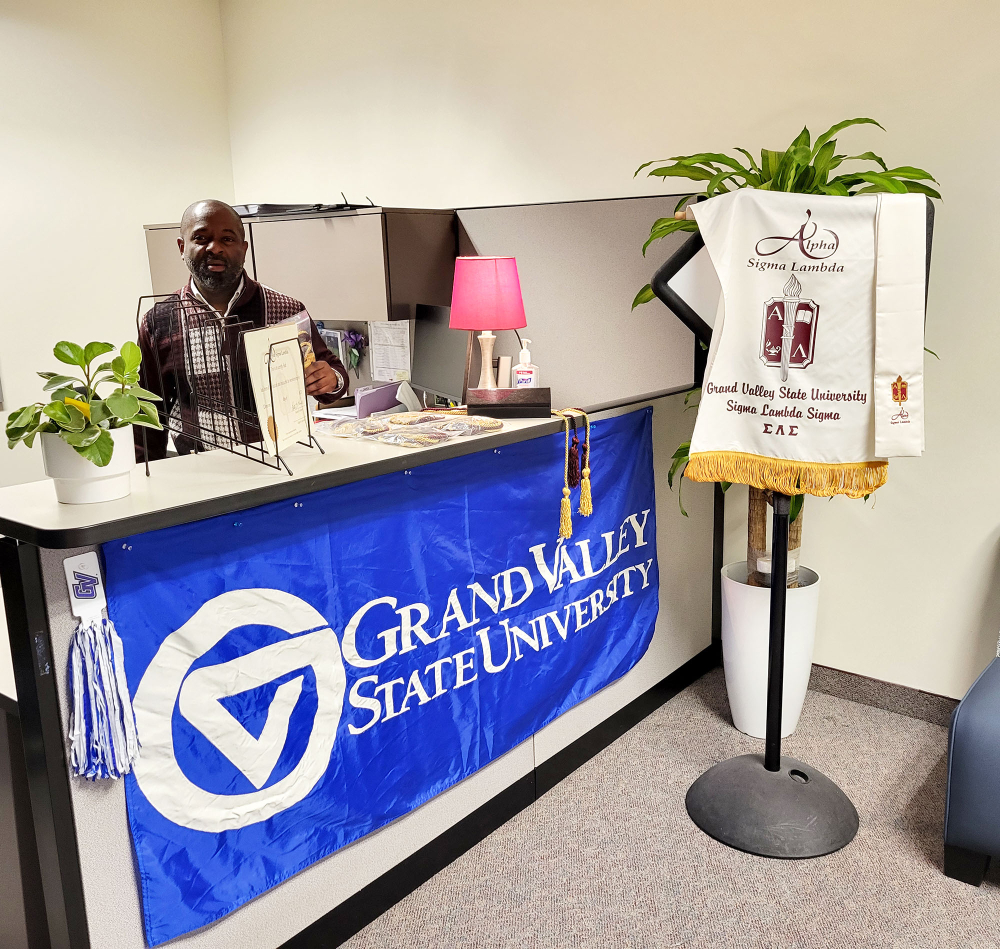 Brian Lassiter stands behind desk, banner for honor society next to GVSU flag pinned to outside of desk