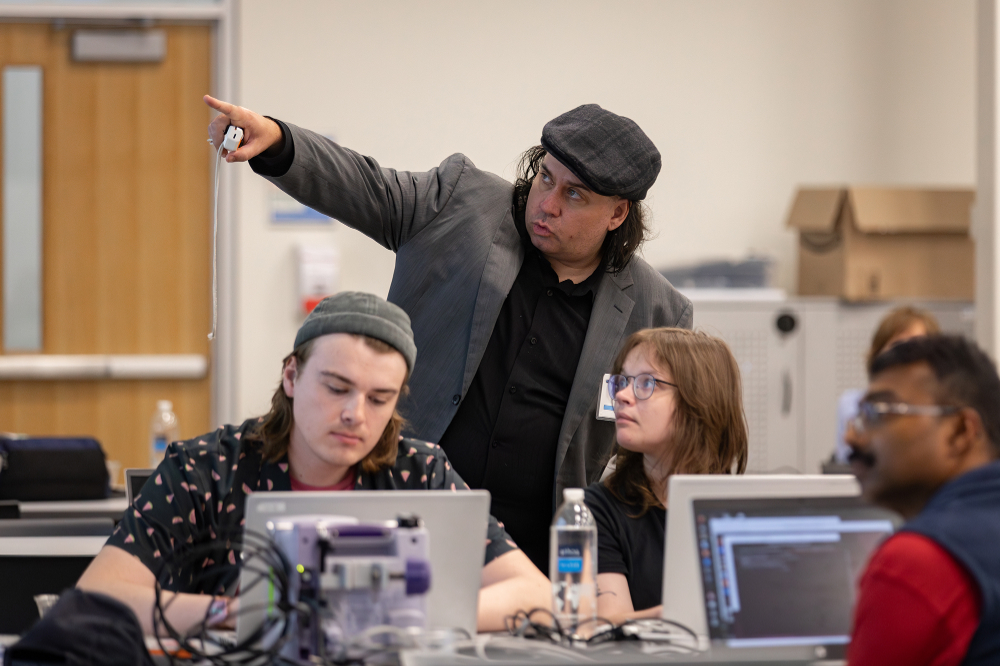 instructor standing and pointing, three students on laptops looking where instructor, in hat and suit jacket, is pointing