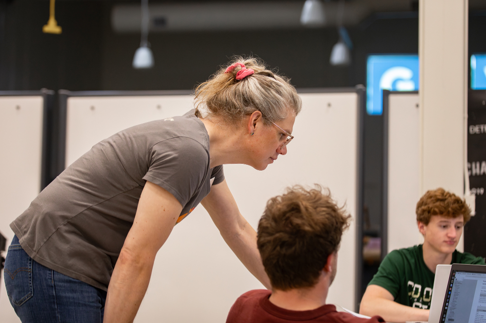 Erin Cornelius stands to look over the shoulder of a student on a laptop. Another student with red hair and green t-shirt at right