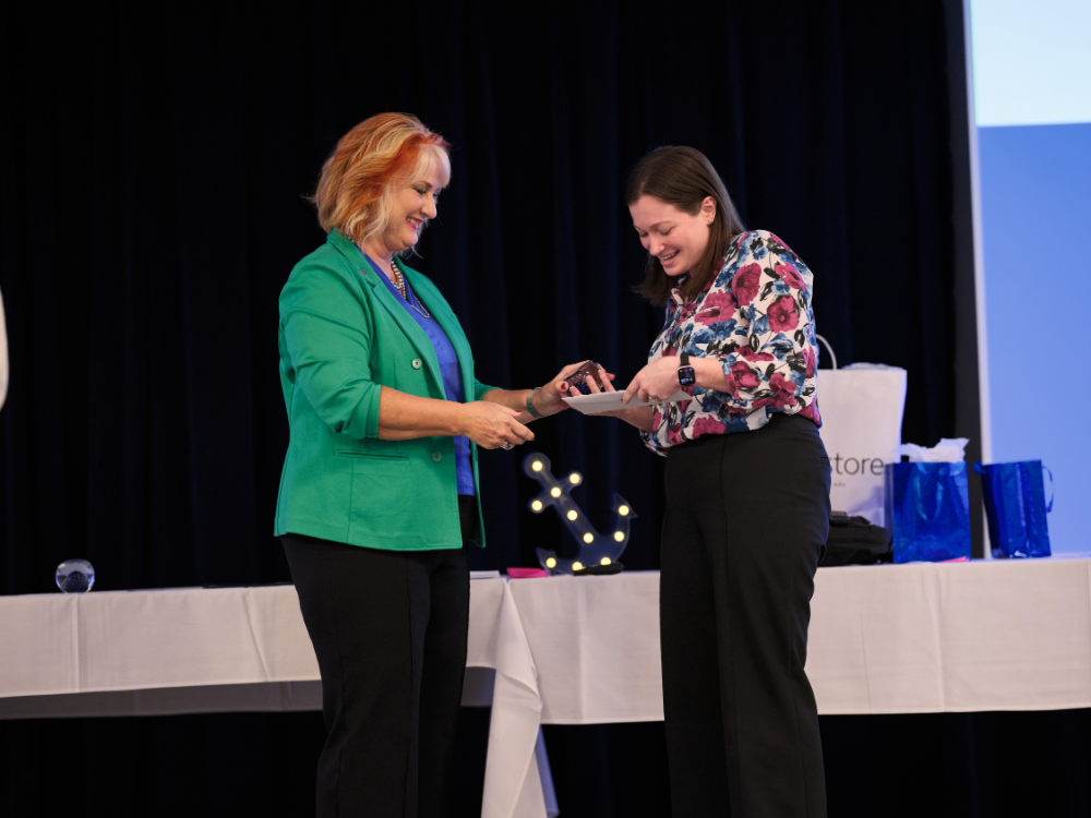 woman in green jacket on left presents award to woman in floral shirt on stage