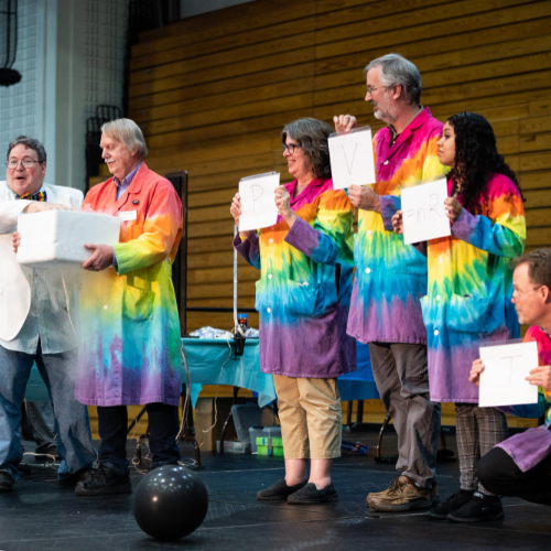 large group on stage holding signs, man at end talks about a box held by another man in a rainbow labcoat