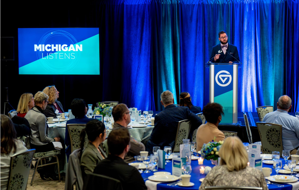Kyle Kooyers at the podium on right, projection screen Michigan Listens in blue and green at left, people seated at tables in front