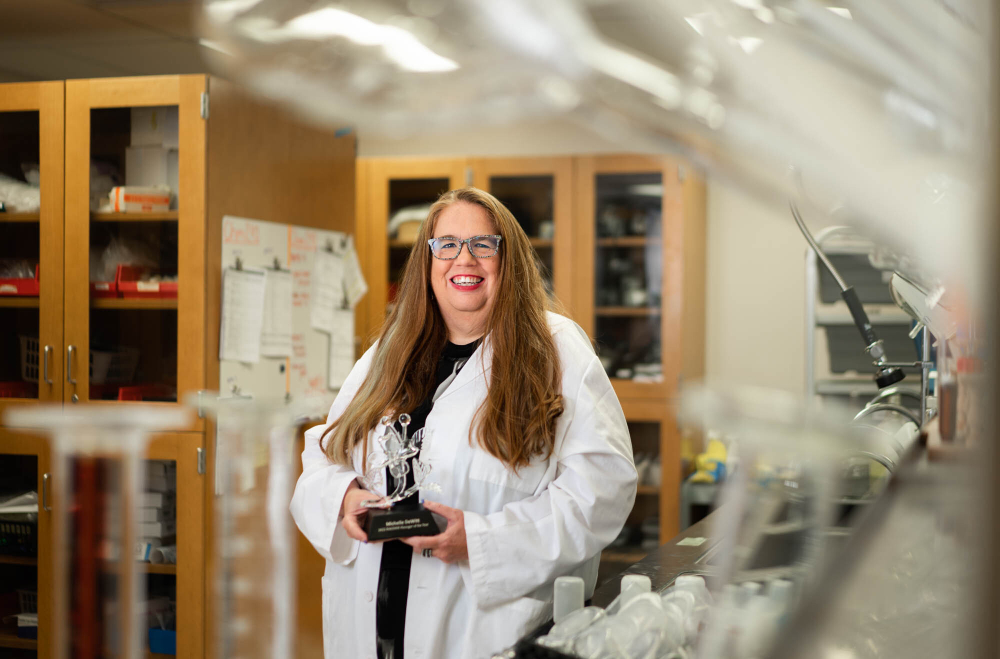 Michelle DeWitt in a white lab coat holds a trophy in a chemistry lab room, the foreground of a piece of clear equipment frames her on the right side