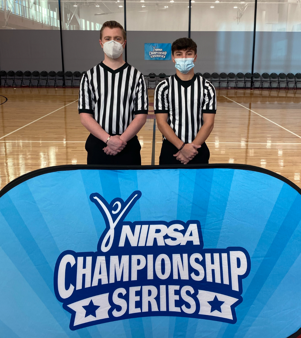two students in referee shirts and face masks stand on a basketball court behind a sign, NIRSA Championship Series