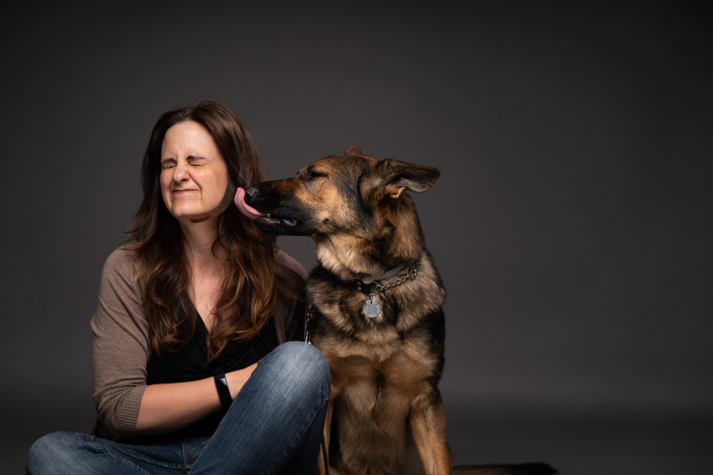 German shepherd licks the face of Jill Hinton Wolfe as she sits on the floor.