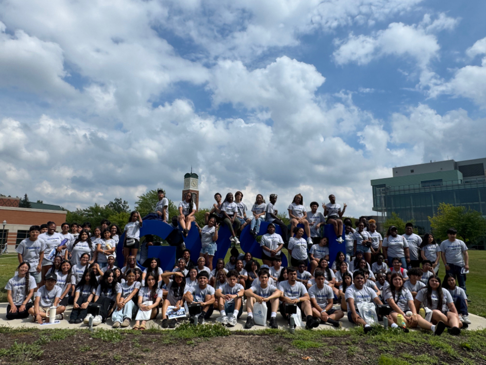 large group of students sitting on or near the GVSU letters, blue sky with lots of clouds