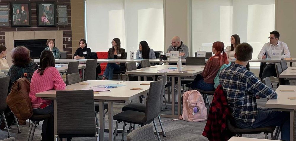 people seated at long table by fireplace look over groups of students gathered and seated at square tables