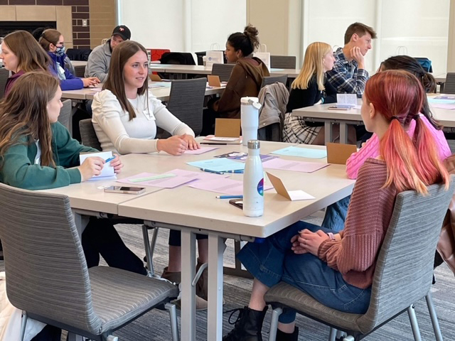 students seated at square tables chat, colorful papers on tables and water bottles