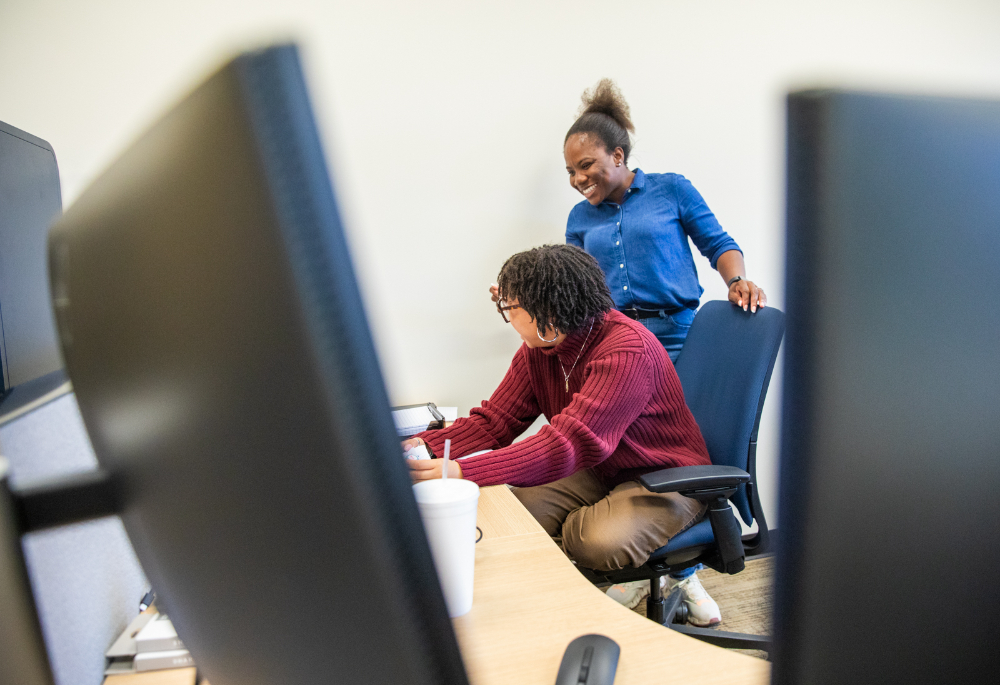two people in background, one standing and one seated, computer monitors frame them from the front