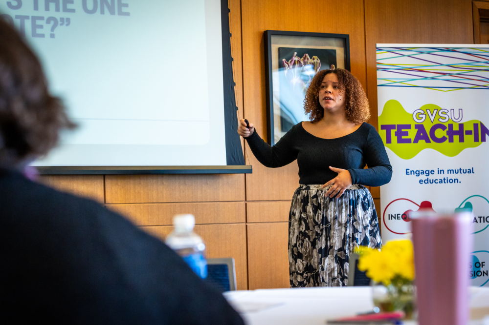 woman pointing at screen at front of classroom, Teach-In poster behind her