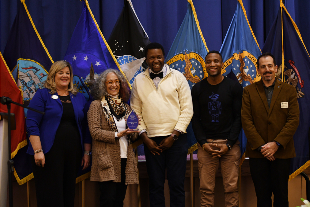 five people standing on stage in front of flags of military branches