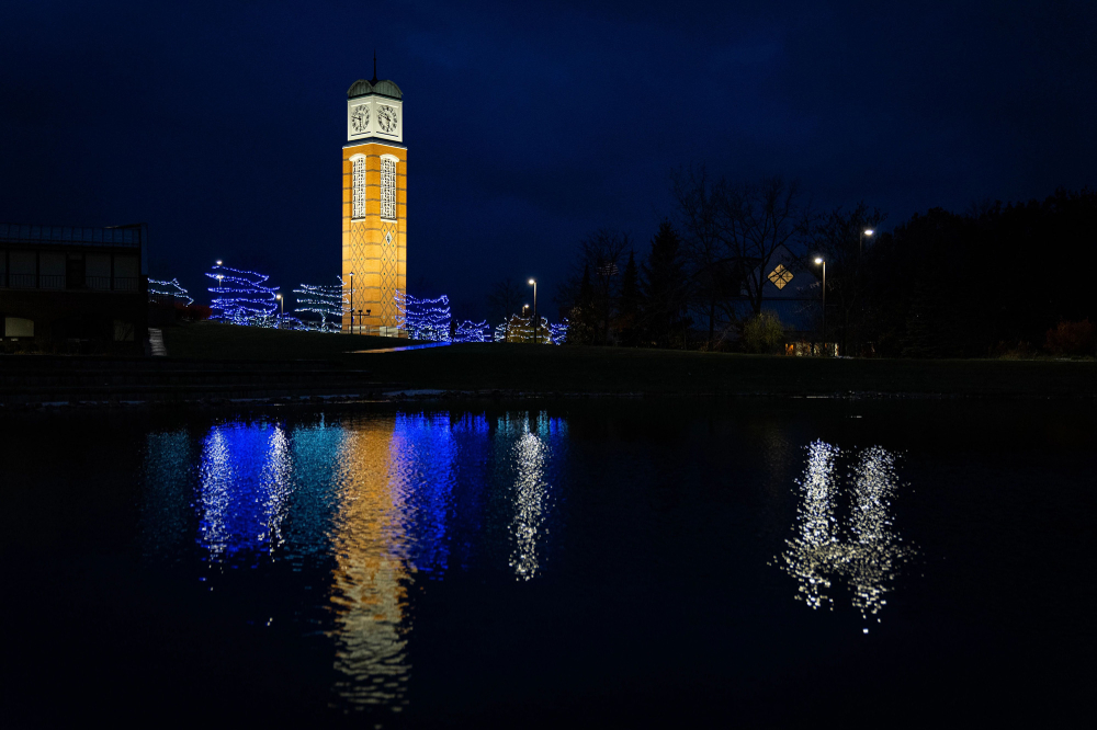 night photo of carillon tower lit up with surrounding trees lit with blue lights; all reflect on Zumberge Pond