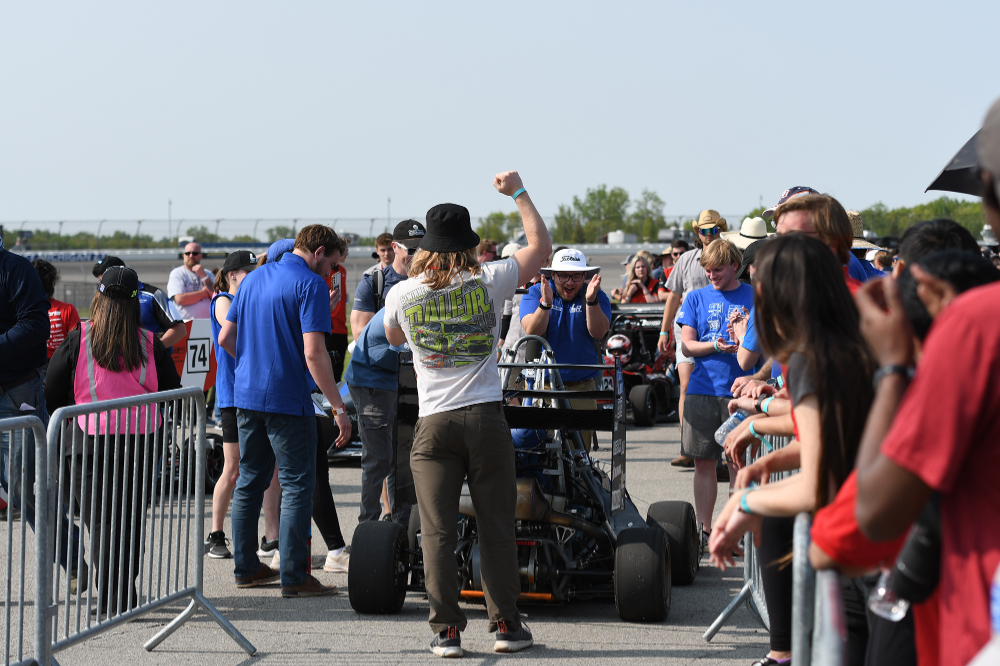 a crowd gathers around an open-wheel race car at the Michigan International Speedway; crew members for Laker Racing are wearing blue shirts