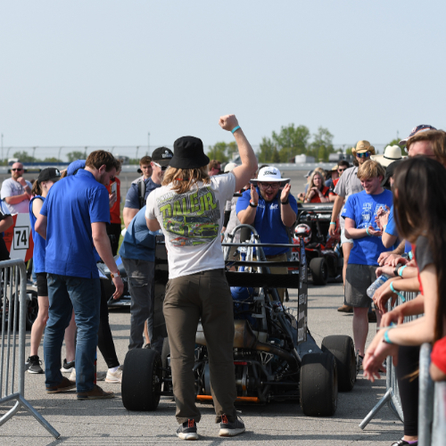 a crowd gathers around an open-wheel race car at the Michigan International Speedway; crew members for Laker Racing are wearing blue shirts