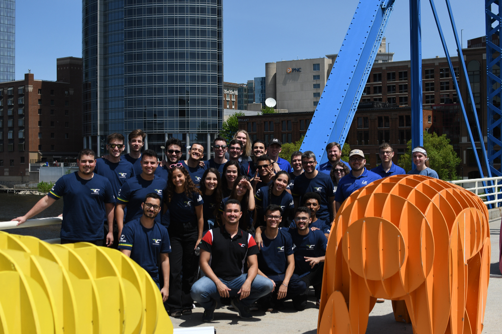 four rows of people stand and kneel on the Blue Bridge in Grand Rapids, behind an art installation of colorful animals and in front of the JW Marriott
