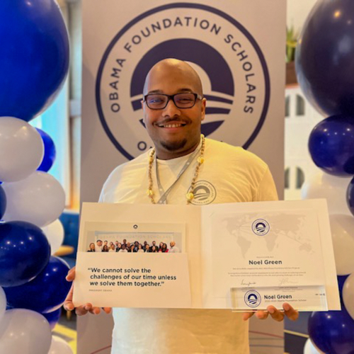 man holds certificate folder in front of background of white and blue balloons and the Obama Foundation Scholars program vertical banner
