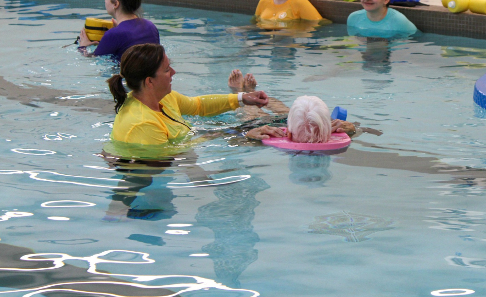 pool with a person in a yellow tshirt helping an older woman float using a pink floating behind her head