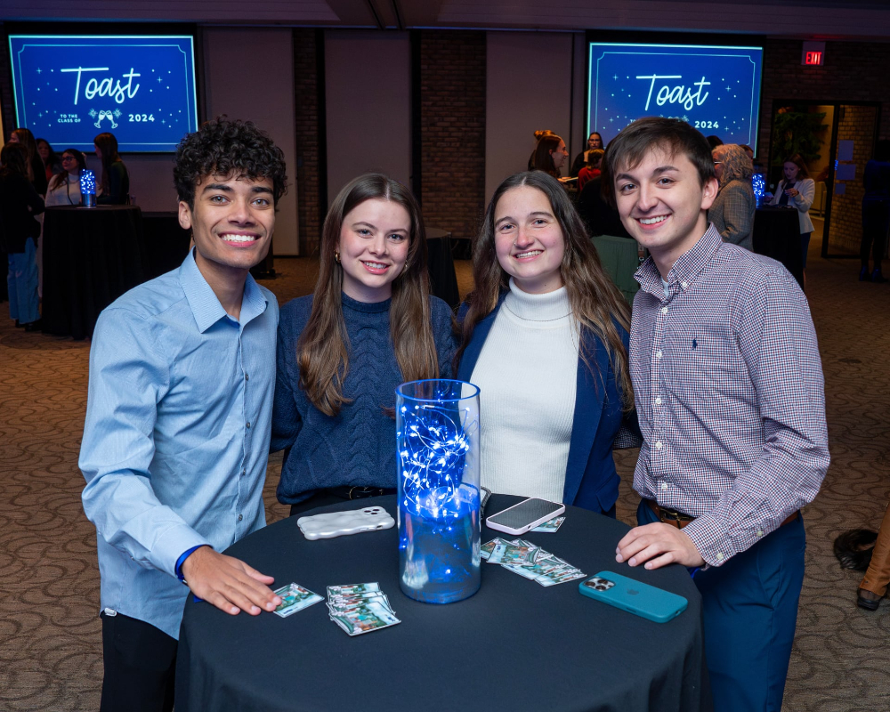 four students standing behind tall table, screens in back read Toast