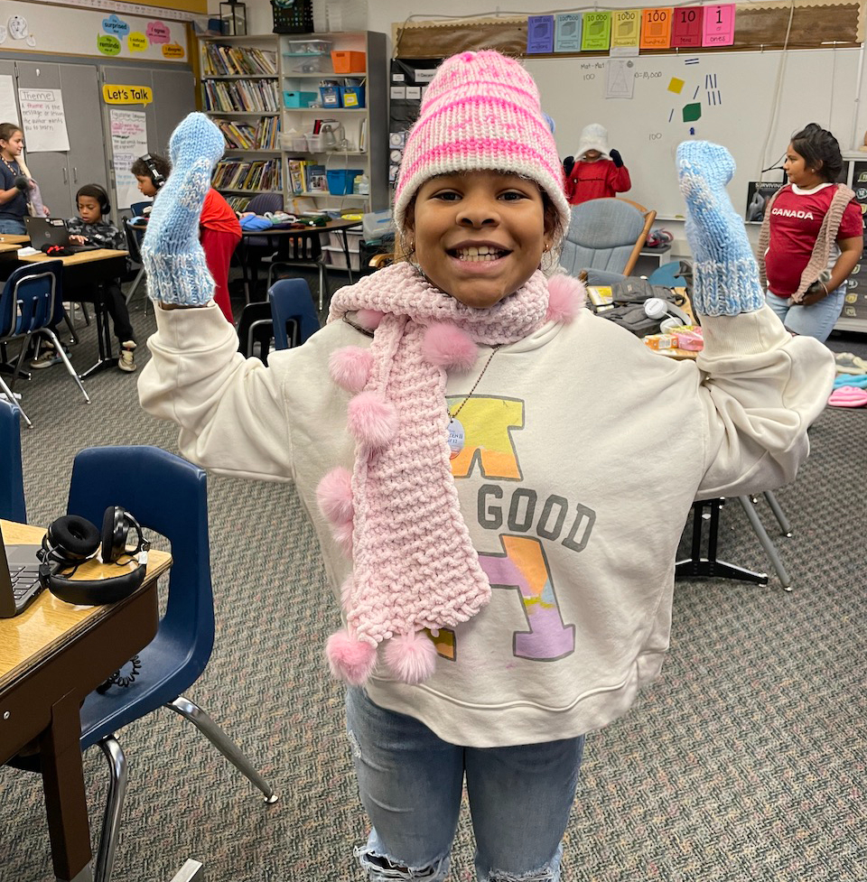 an elementary student shows off knitted mittens, hat and scarf inside a classroom