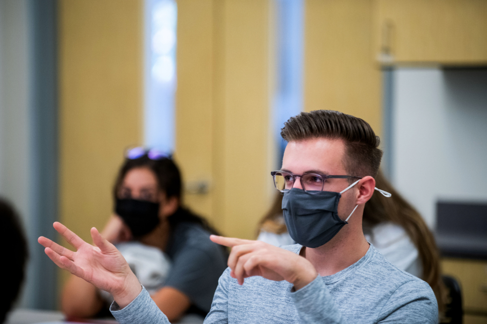 student in face mask speaks from his seat in a room and gestures with hands