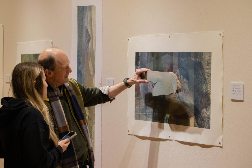 two people looking at artwork on a wall in an exhibit; man is holding piece of wood next to artwork