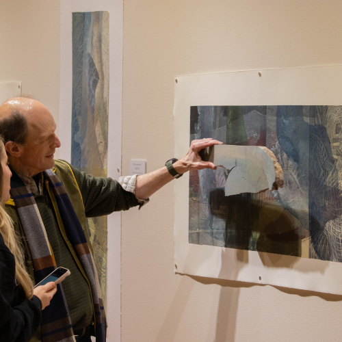 two people looking at artwork on a wall in an exhibit; man is holding piece of wood next to artwork