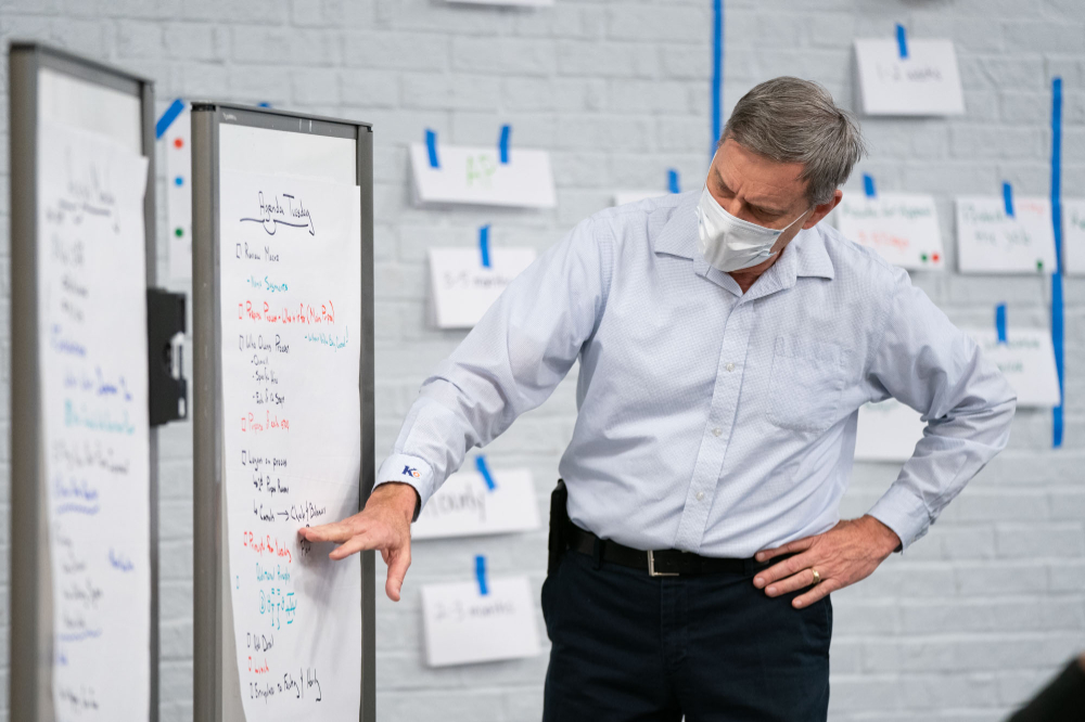 man standing pointing at one of two white boards, a wall full of papers taped to it is behind him