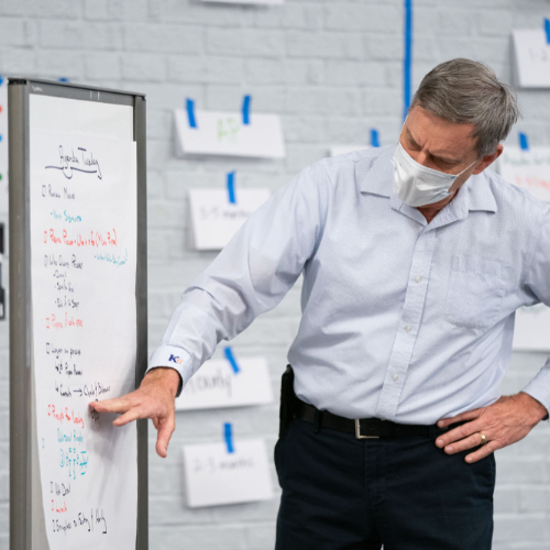 man standing pointing at one of two white boards, a wall full of papers taped to it is behind him