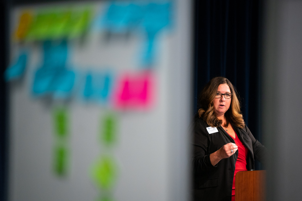behind a large whiteboard filled with colorful post it notes, a woman stands at a podium.