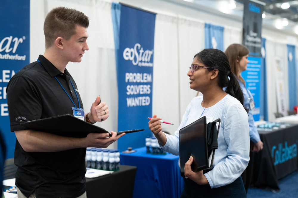 two people talk at career fair, woman in white shirt, man in black polo shirt on left, company banners hanging on white partitions behind tables