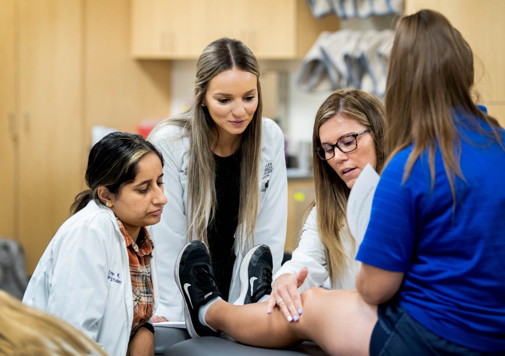 student in blue shirt and shorts sits on medical table while three students in white coats look at her knee during a simulation exercise