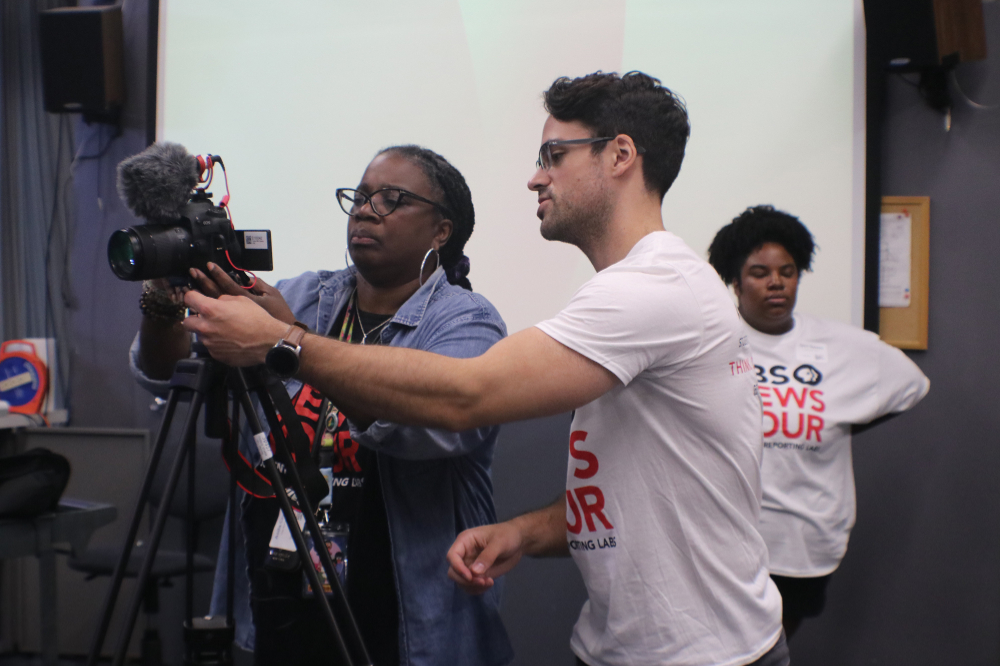 two people behind a video camera, one wearing white PBS news hour tshirt, other person in denim shirt