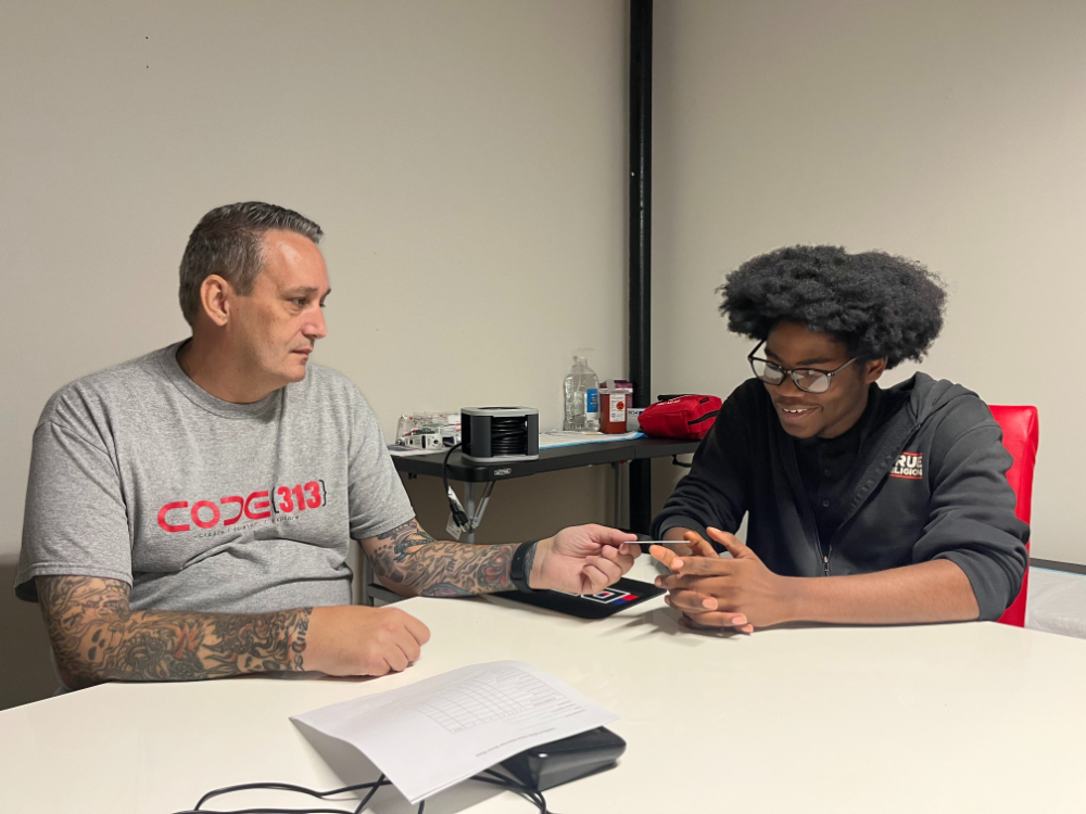 two people seated at table, man on left has CODE313 tshirt on