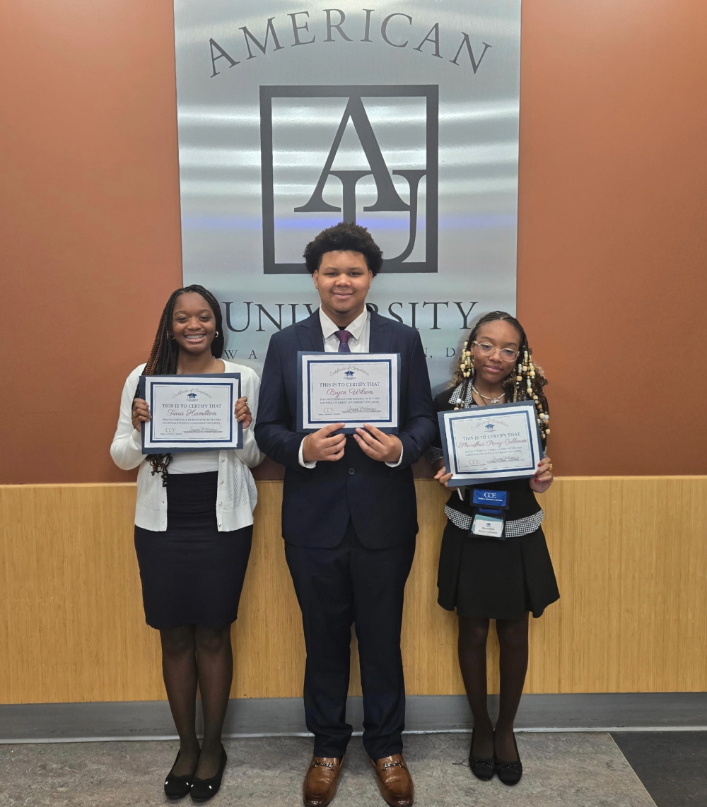 three students holding certificates in front of an American University sign inside a building
