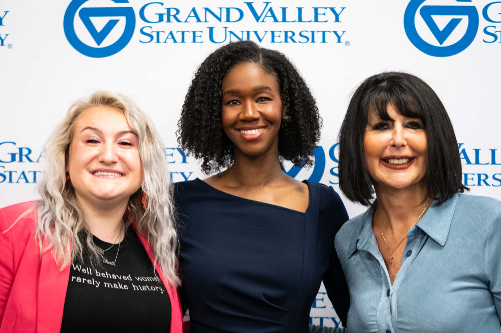 left to right are Student Senate President Faith Kidd, Justice Kyra Harris Bolden and President Mantella against a photo backdrop with the GVSU wordmark
