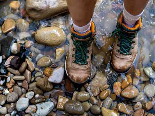 Closeup of a persons hiking boots standing in shallow water on river rocks