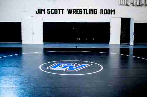 Black text on the white stone wall reads 'Jim Scott Wrestling Room.' Black doors line the wall and the black padded floor has the athletics GV logo stenciled on it. 