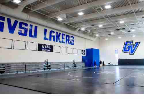 A large room with a black padded floor and white walls. GVSU Lakers and the GV athletics logo are stenciled on the walls. At the back of the room three people are gathered, speaking.
