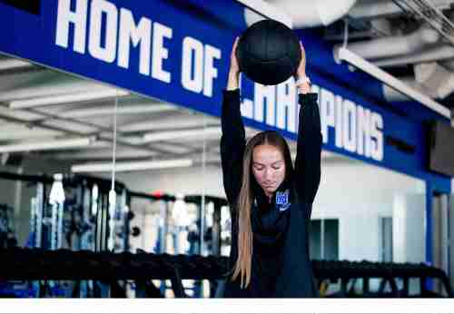 A student lifts a weighted slam ball into the air. In the background is a workout space with mirror and 'Home of the Champions' is written on the wall. 